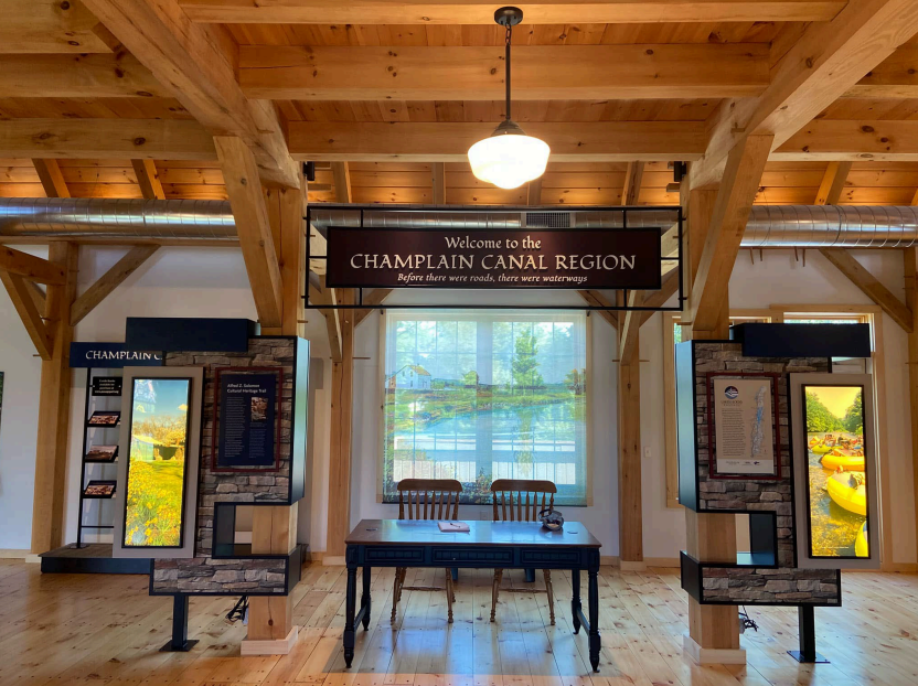 An interior space with a rustic, wooden design featuring exposed beam and high ceiling. There’s a sign that reads “Welcome to the Chaplain Canal Region” with a tagline “Before there were roads, there were waterways”. Below the sign is a table and two chai