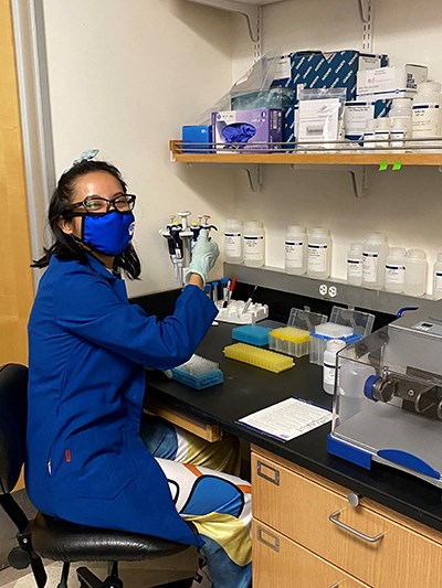 A woman in a blue lab coat and mask works at a laboratory bench