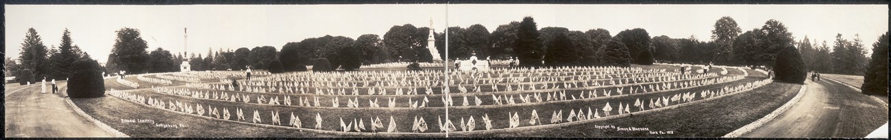 Panorama of cemetery with rows of burials and small flags curving outward from a tall monument