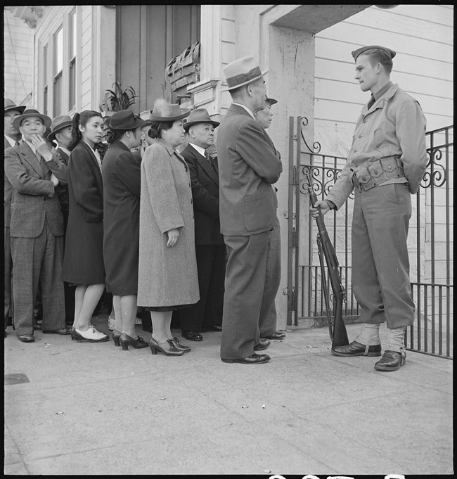 A soldier with a gun stands in front of a low gate facing a line of Japanese American adults