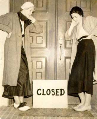 Two stylish women lean against closed door, contemplating “closed sign” at their feet.
