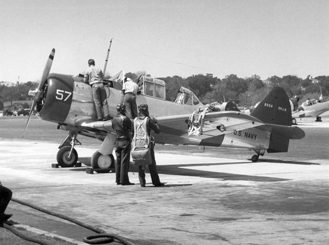 Black and white photo of four men, two in flight suits around a propeller plane