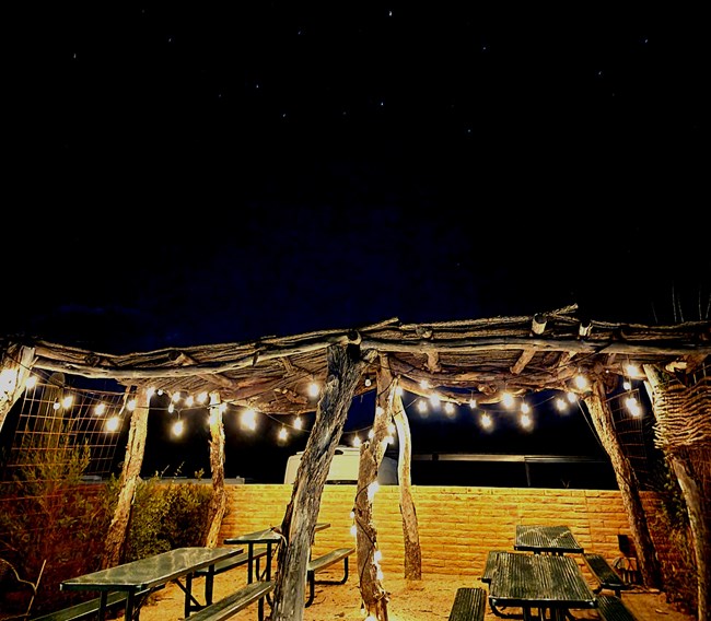 A ramada with string lights hanging down illuminate picnic tables below a night sky.