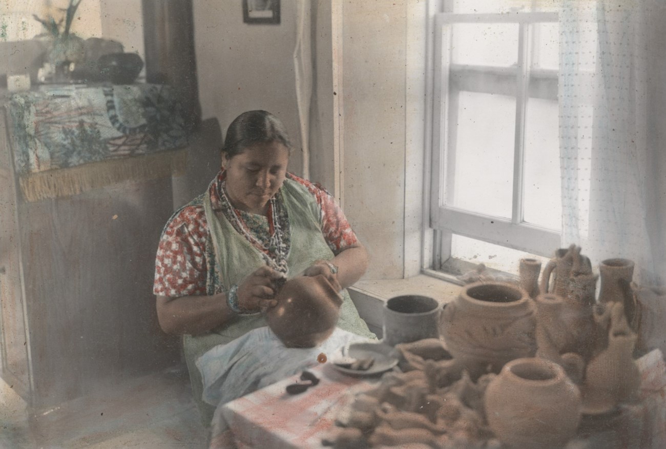 Legoria Tafoya sits at table covered in pots and animals, painting a pot in her lap.