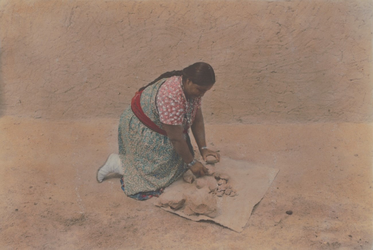Legoria Tafoya kneels on the ground breaking up clay clumps.