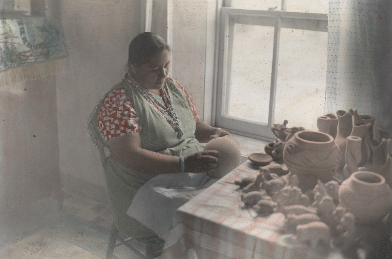 Legoria Tafoya sits in front of a window at a kitchen table covered in pots and animal figures as she sands a round pot.