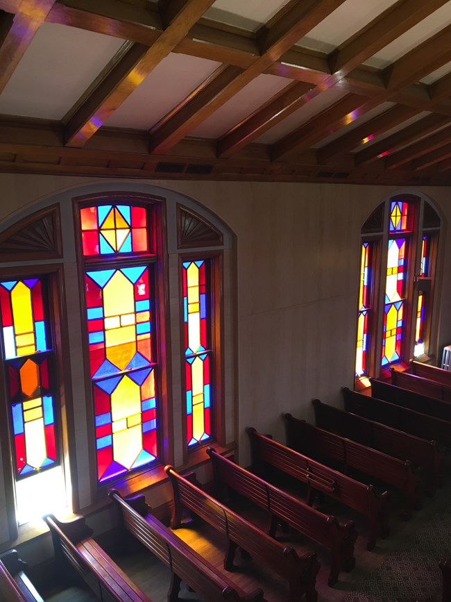 The pews of the sanctuary space of the synagogue against the tripartite geometric stained glass as seen from the organ loft.