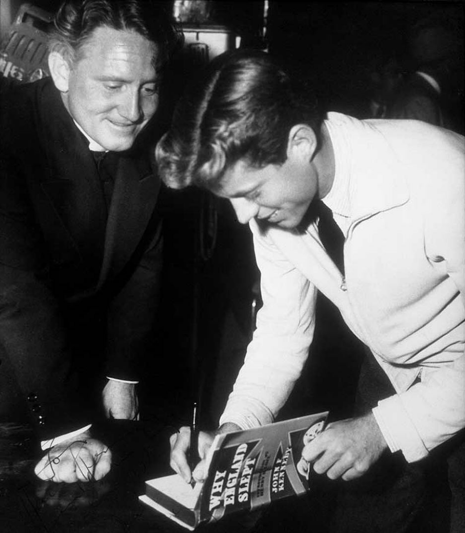 A black and white photo of John F. Kennedy, wearing a white jacket, signing the inside the book ‘Why England Slept’ as Spencer Tracy watches in a black suit.