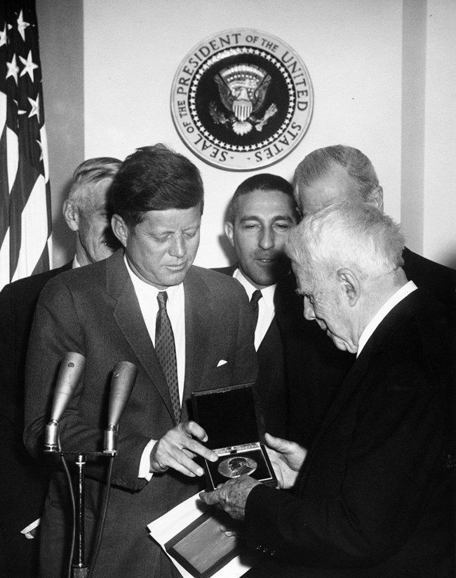 A black and white photo of President Kennedy handing a medal to a white-haired man in a suit, as other men look on, with a Presidential seal on the wall and an American flag visible in the background. Two microphones are at Kennedy’s right.