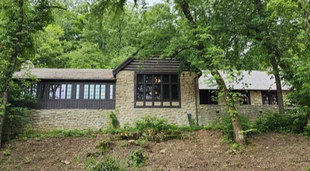 thumbnail_Outlook-fdqmnhb2 A grey stone lodge with dark wood roof and trim. Large windows are visible from the front perscepctive of the building. A lush forest of green trees are surrounding the structure.