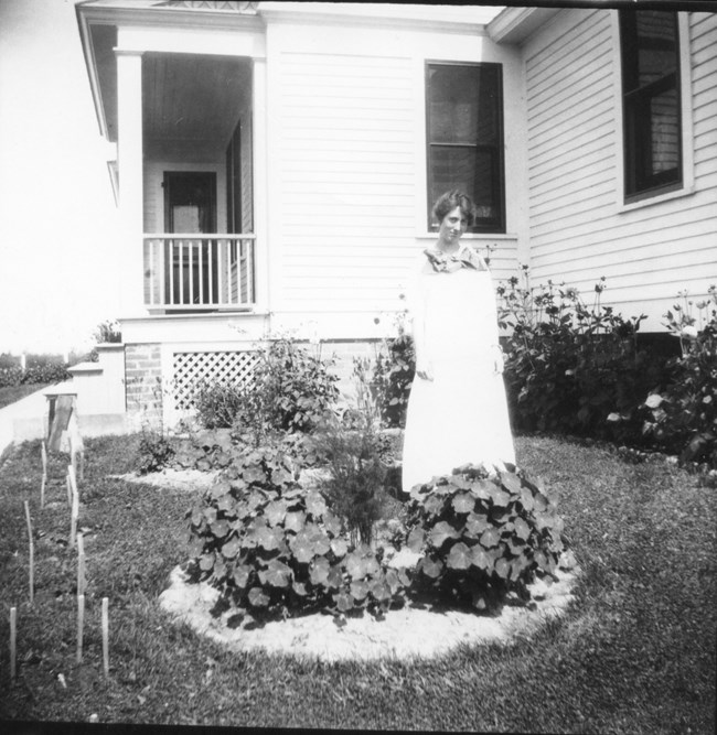A black and white photo of a woman in a white dress standing in front of flowers.