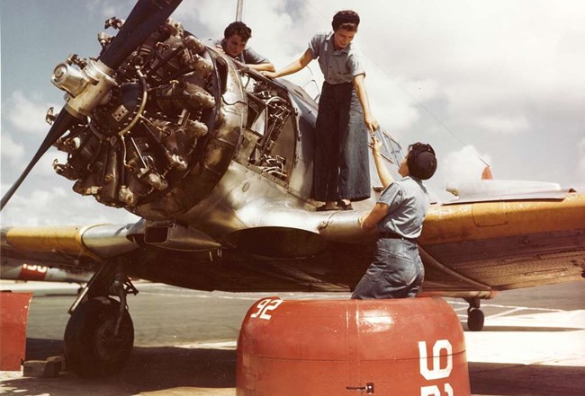 Color photo of three women in denim working on a propeller plane with the engine exposed. One woman hands something to the other two women standing on the wings of the plane