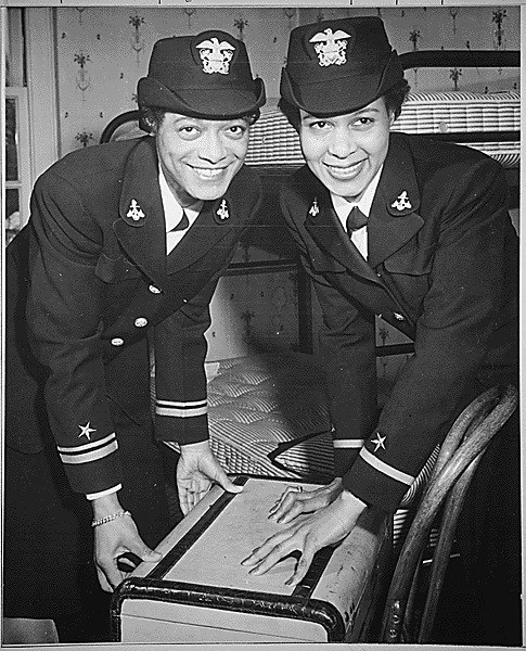 Two African American women in military uniforms smile while bending over a suitcase with bunk beds in the background.