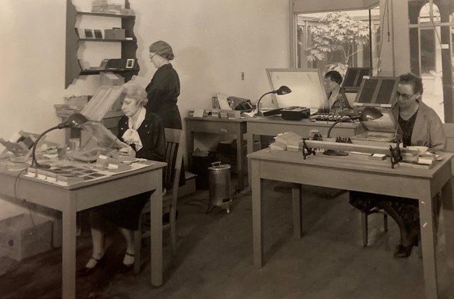 Three women sitting at desks in front of lighted magnifiers. One woman stands at a shelf.