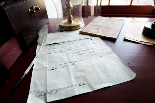 Reproduction historical document with cursive writing in ink on period parchment lies on dark wooden desk, with feather quill to left. Other colonial-era objects adorn desk, including wooden drawer, pamphlets, and glass-encased lamp in background.