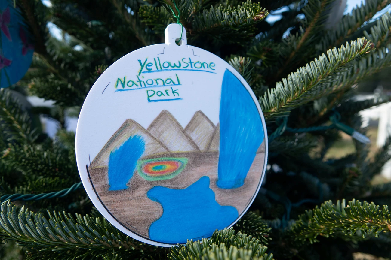 yellowstone national christmas tree 2021 ornament with a drawing of mountains and yellowstone's geyser in the middle. Ornament is hanging from christmas tree