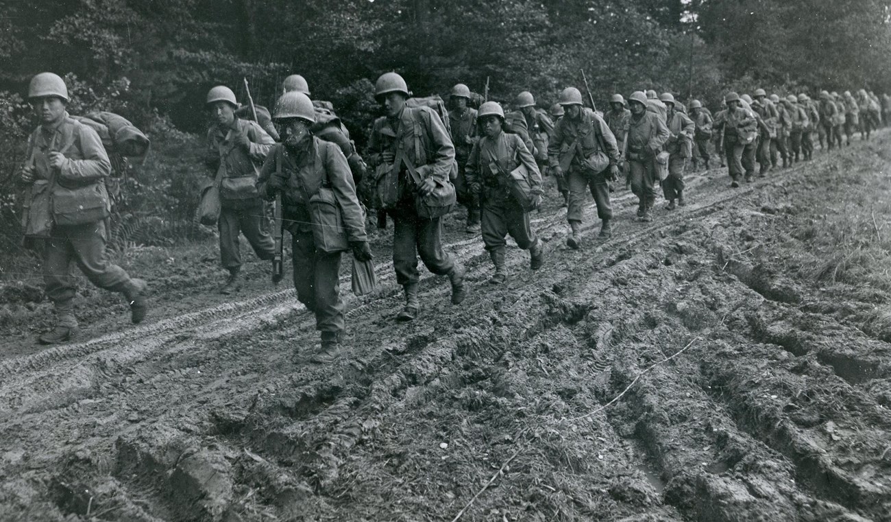 A black and white photo shows a group of soldiers in uniform marching up a muddy path.