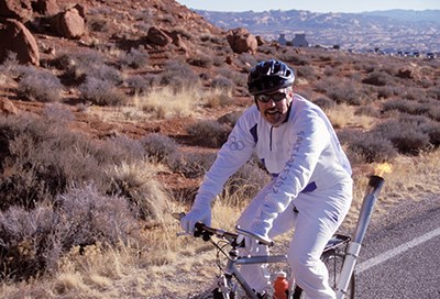lt= a cyclist in white Olympic uniform with a lit torch on the back of the bicycle