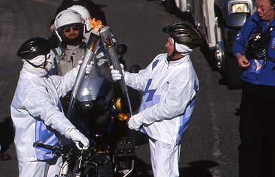 lt= two people wearing white pass the Olympic flame from one torch to another