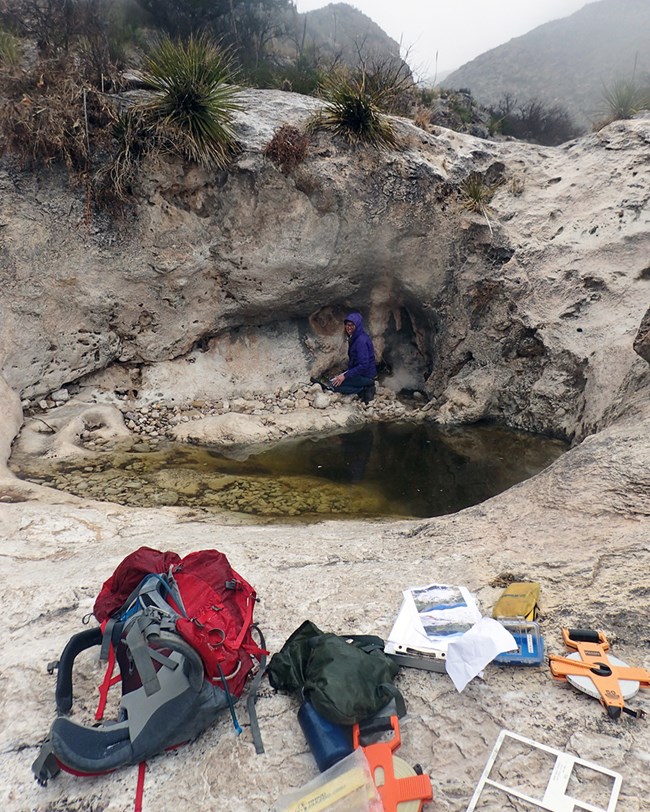 A scientist, backpack, measuring tapes, and clipboard next to a pool of water in a bedrock depression.