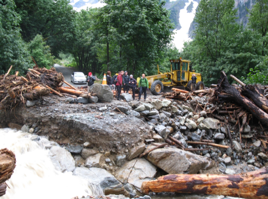 People with heavy equipment looking out over water flowing through a collapsed section of road littered with woody debris