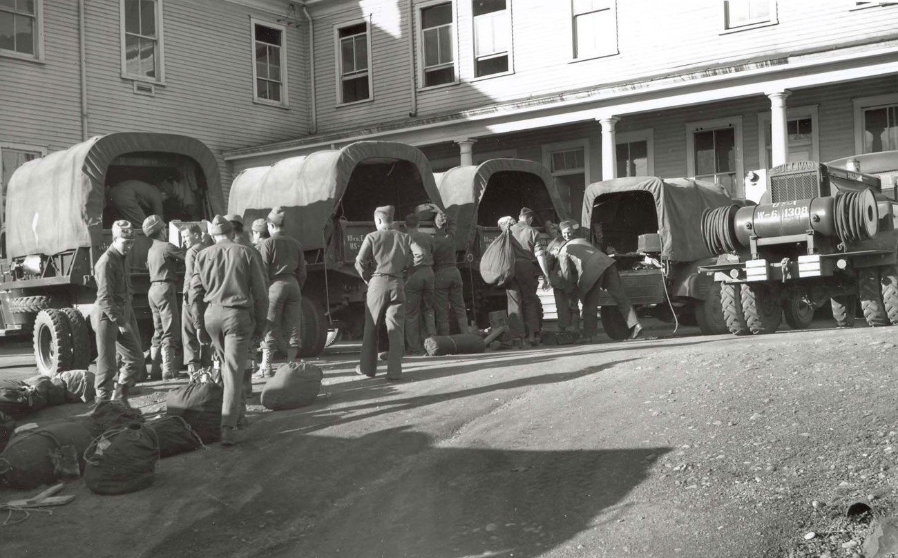 A black and white photo shows soldiers in WW2 uniforms loading supplies into covered trucks. Behind them is a two-story building with a porch.