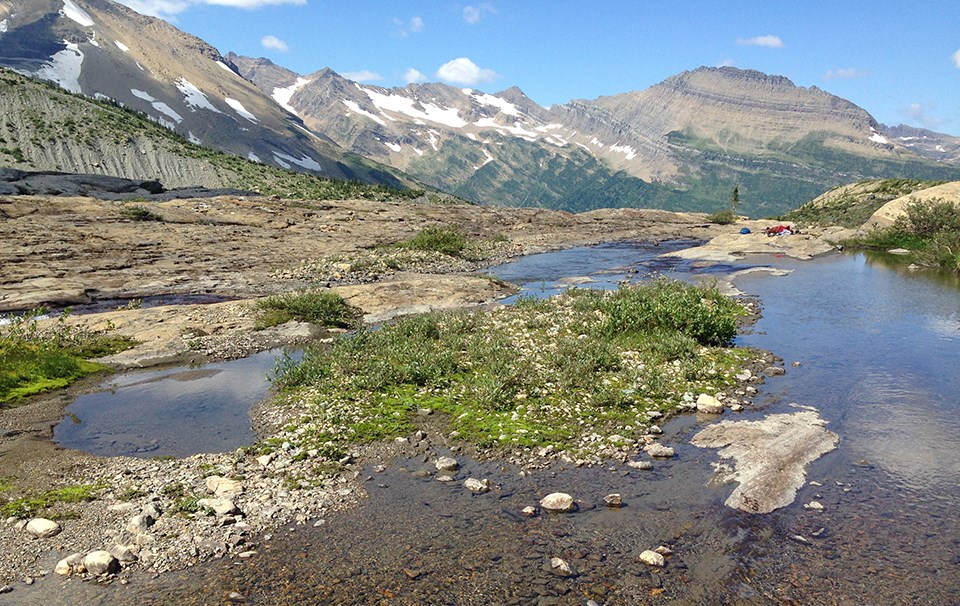 patches of snow on high mountain ridges with stream flowing across rocky landscape and a few riparian shrubs