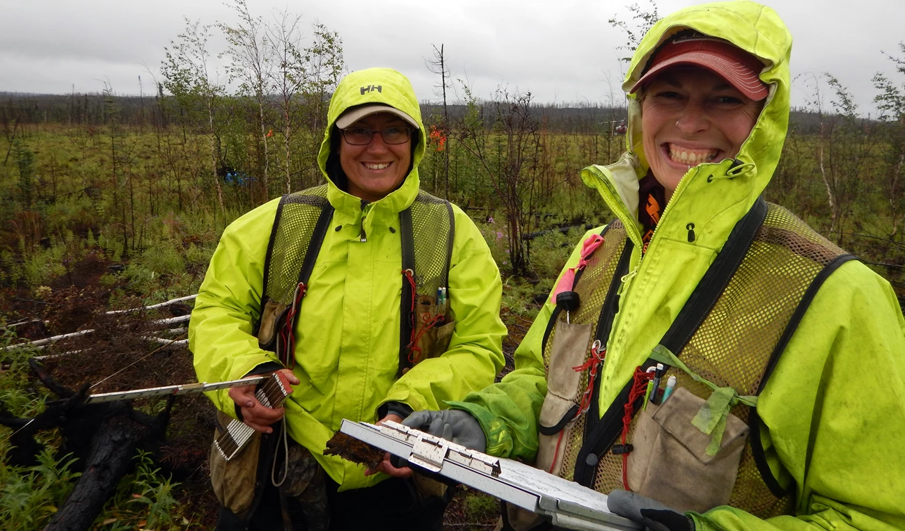 Fire effects monitoring in Denali National Park Two people with neon green rain jackets hold paperwork and a measuring stick.