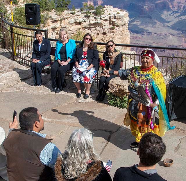 Havasupai elder wearing colorful clothing and holding a rattle and feathers is conducting a welcome ceremony.
