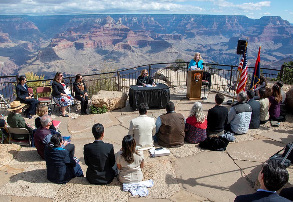 15 people sit in a semicircle in a stone amphitheater. A judge in black robes is sitting at a table with a black tablecloth. A Woman dressed in blue is behind a podium and addressing the group. In the distance, a colorful landscape of peaks and cliffs.