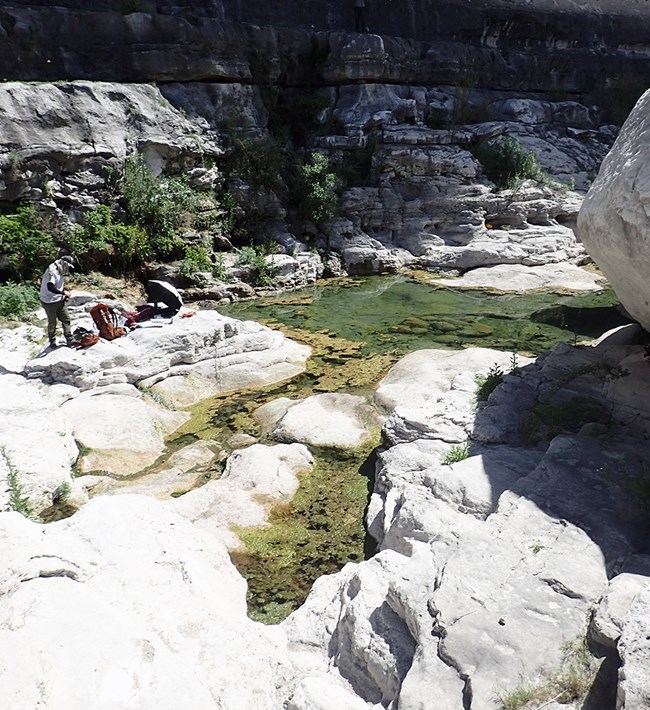 A pool of greenish water in front of a rock face with large boulders around it.