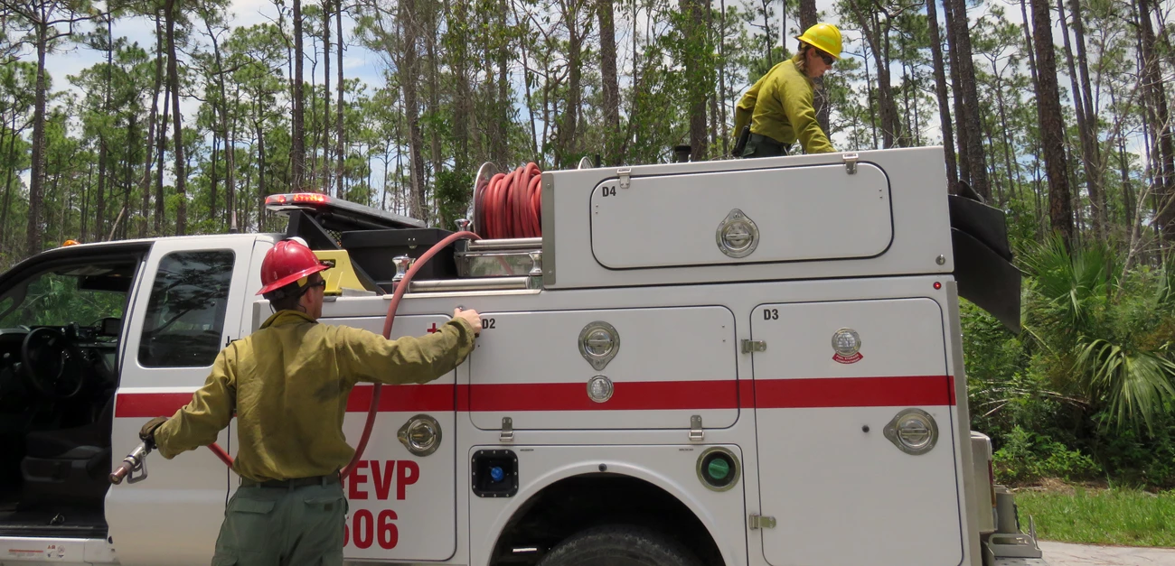 Engine crewmembers A firefighter pulls a hose down from an engine while another firefighter on top of the engine leans over.