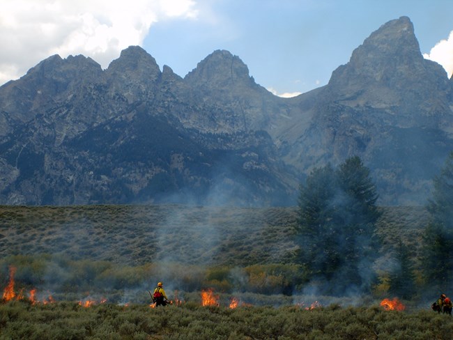 Firefighters ignite grasses and sagebrush with the Grand Tetons looming.