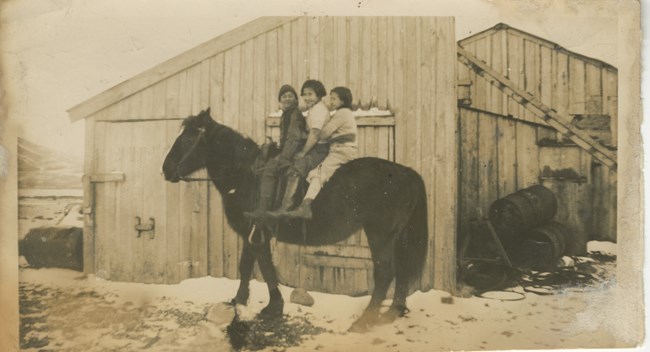 black and white photo of three children sitting on a horse, in front of a building