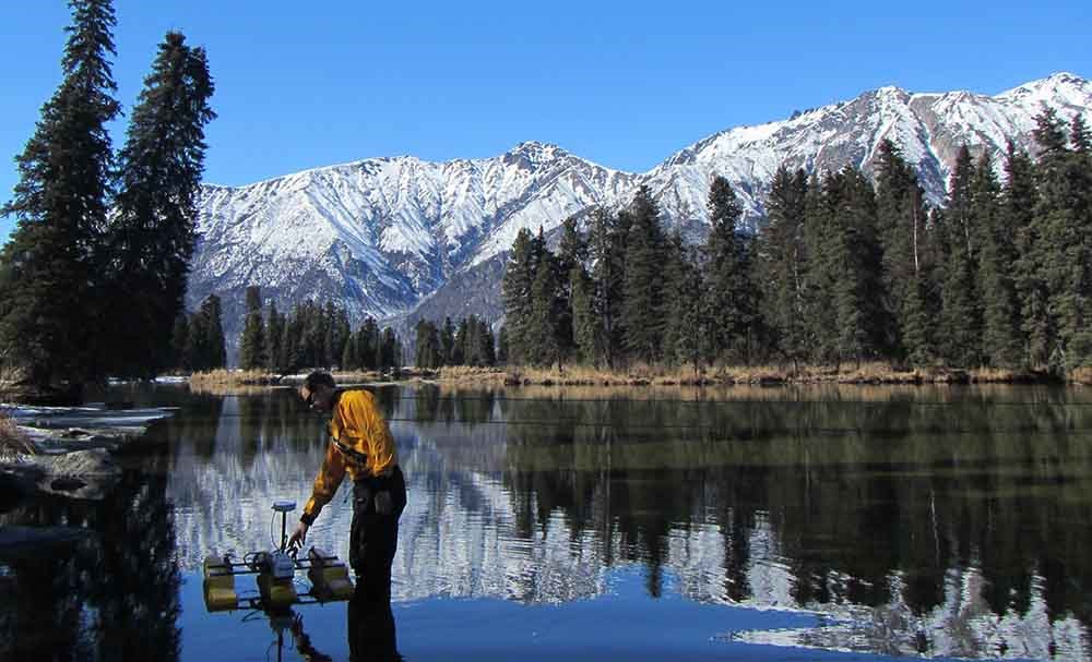 A researcher collects water discharge measurements at a lake with mountains in the background.