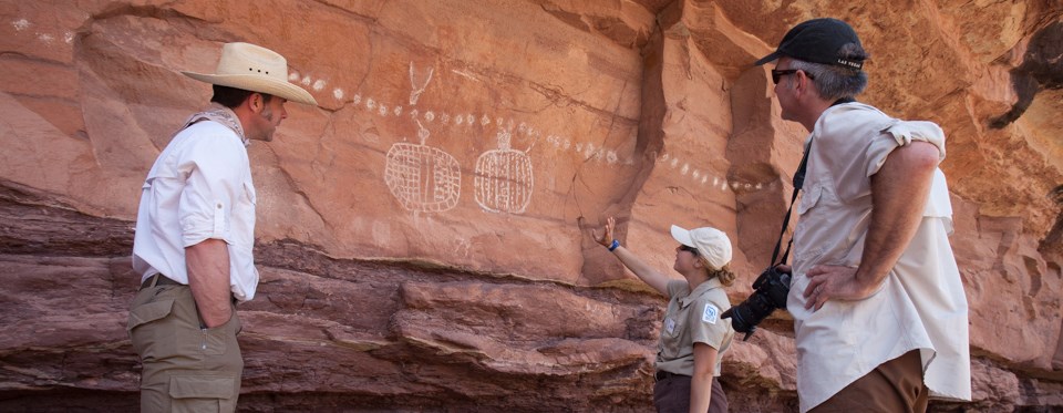 three people stand in front of a rock wall with white painted figures on it