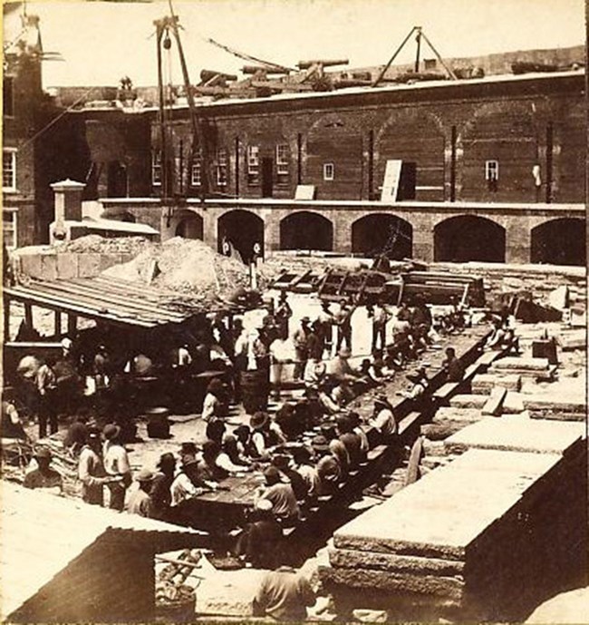 Left Face Being Repaired Photograph of Fort Sumter's left wall being repaired. Workers seated along long table on parade ground. Temporary kitchen on parade ground with long line of men.