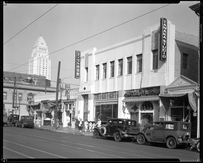 A black and white photo shows a street with several storefronts. There are four 1920s automobiles parked along the street. Four girls and one man stand outside of the storefront, wearing a mix of Western and Japanese clothes.