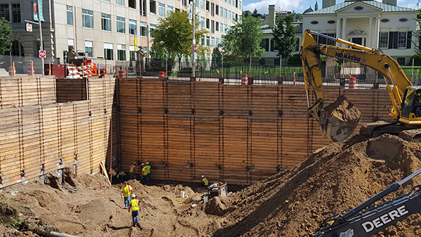 Construction workers use machinery to dig a large hole. The Taft Museum of Art is in the background.