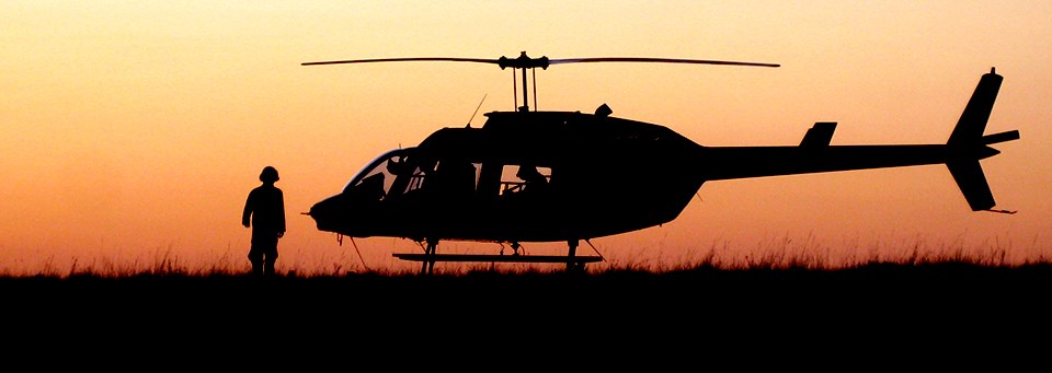 A helicopter sits on the ground as a person in a flight suit walks in front of it during sunset.