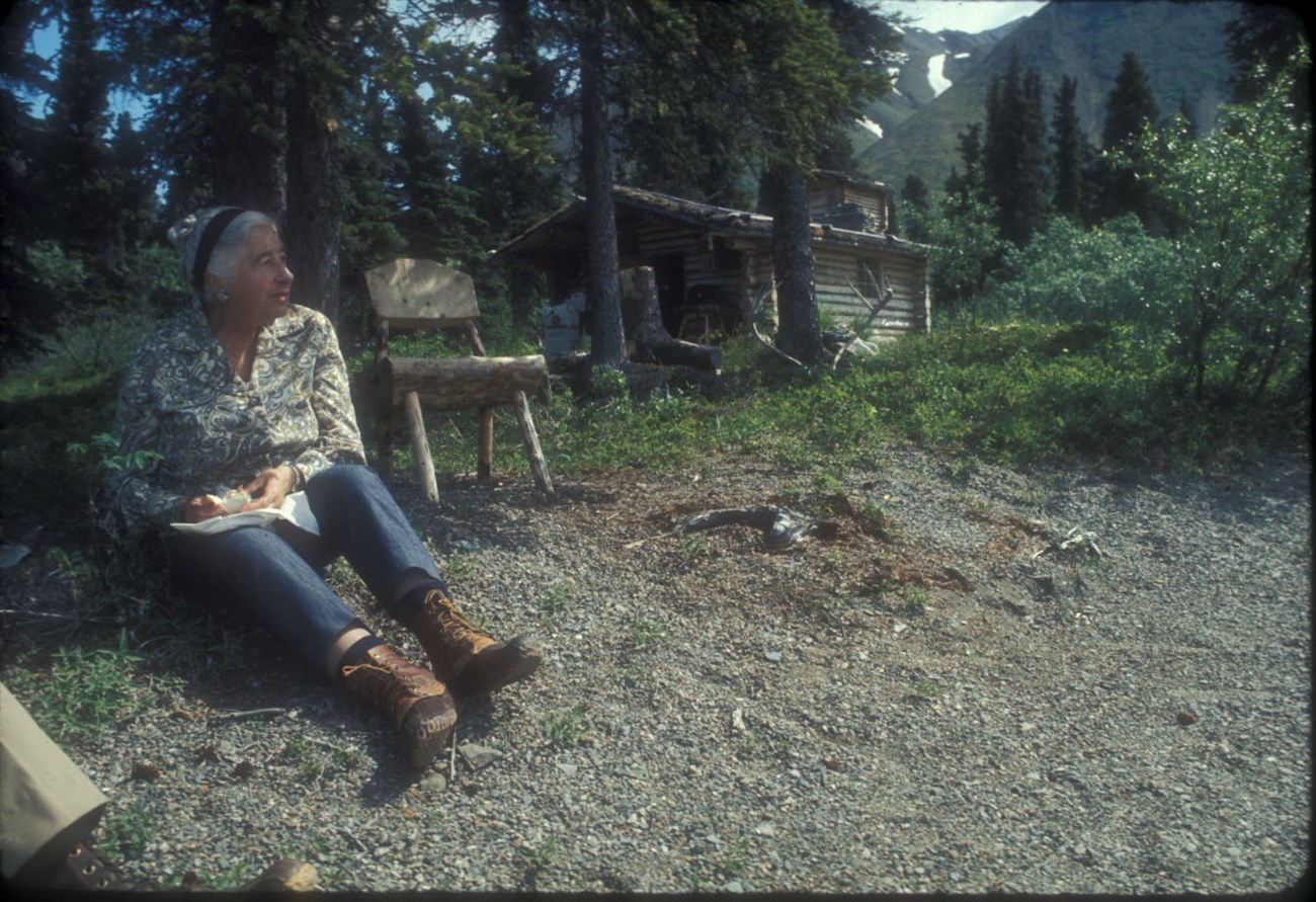 Image of woman sitting on a beach with cabin and chair in background.