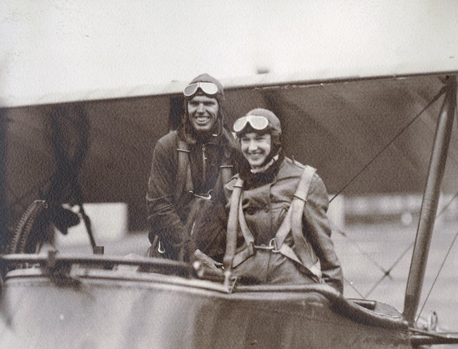 black and white photo of a man and woman wearing flight suits posing in a biplane