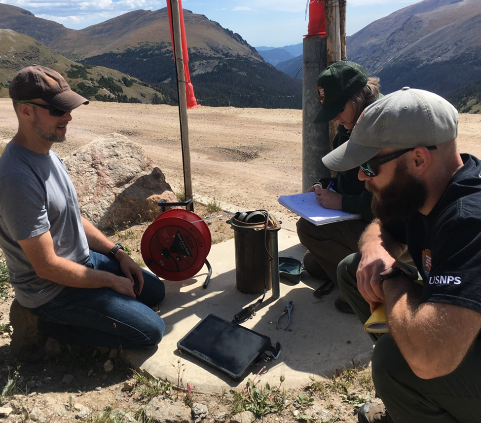 Three people kneel around some equipment beside a dirt road with a view of the mountains.