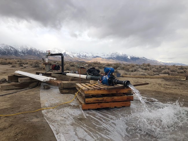 Water gushes out of some equipment connected to a pipe in front of snow-capped mountains.