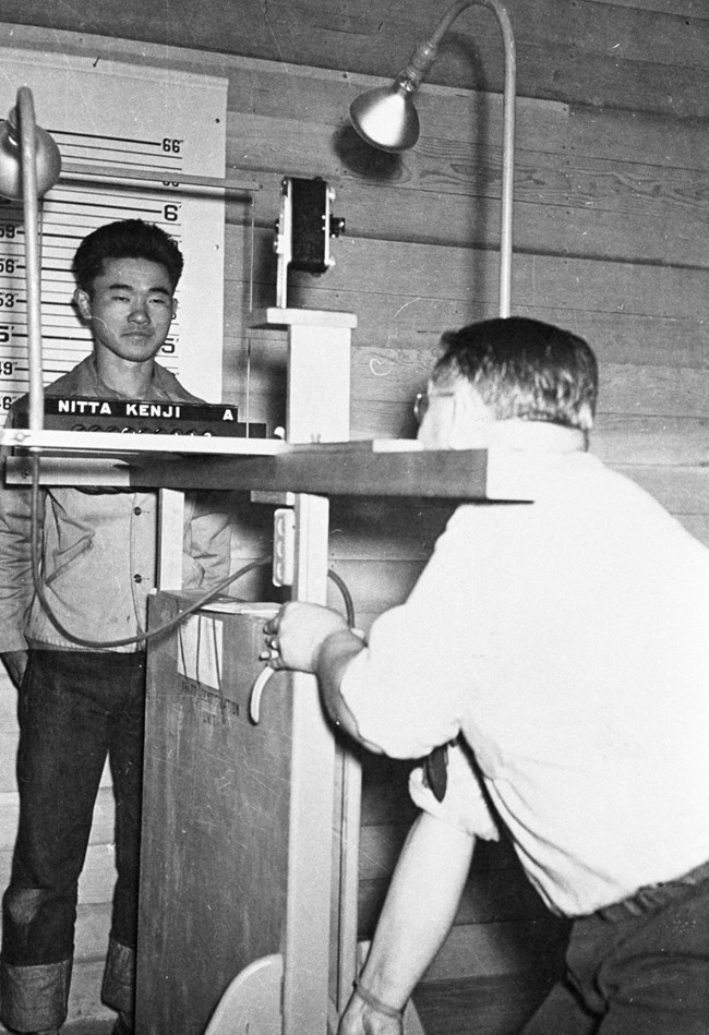 A black and white photo shows a young man being photographed against a wall measuring his height. The nameplate below his face reads "Nitta, Kenji A."