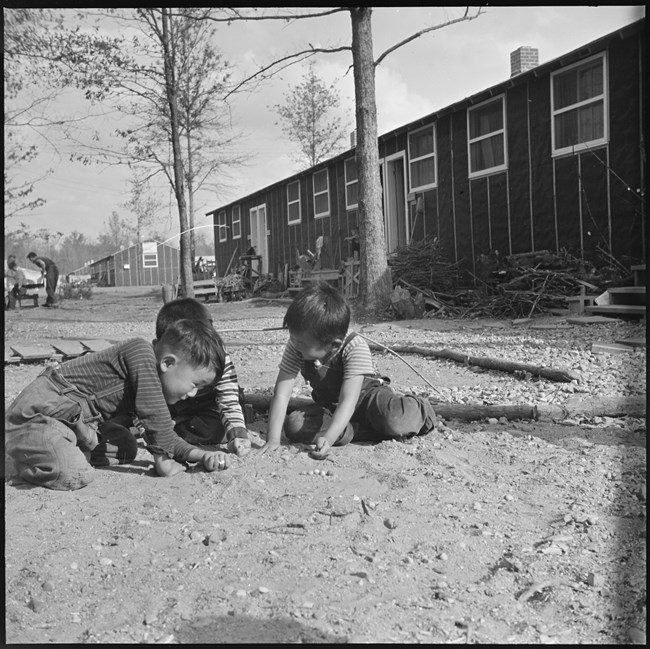 A black and white photo shows three young boys in overalls playing with marbles in the dirt. Behind them is a row of barracks with tar paper walls and white windows.