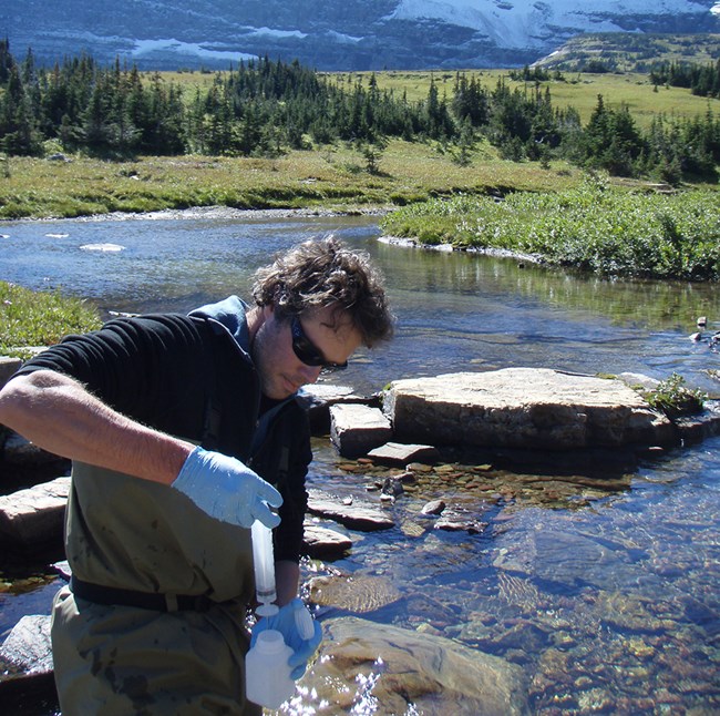 technician wearing plastic gloves and adding a water sample to collection bottle, with stream, forest, and mountains in the background