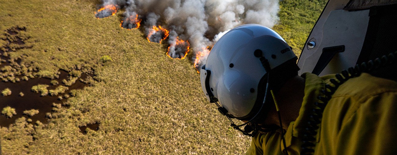 A person in a flight helmet leans out of a helicopter looking at fire burning on the ground below.
