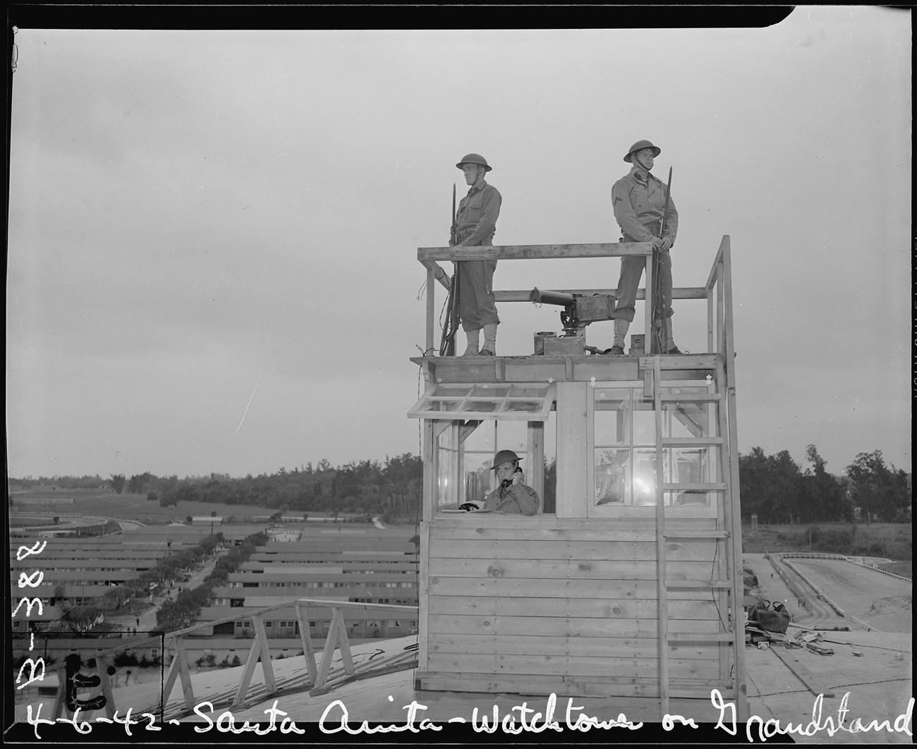 A black and white photo shows a raised wooden guardtower. Two soldiers in uniform with bayonets stand on top of the tower. Another soldier stands inside of the tower holding a black corded phone to his ear.