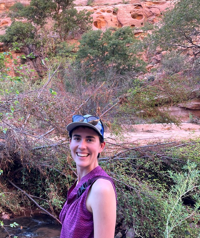 woman with short hair wearing ball cap and pink shirt with trees and canyon in the background